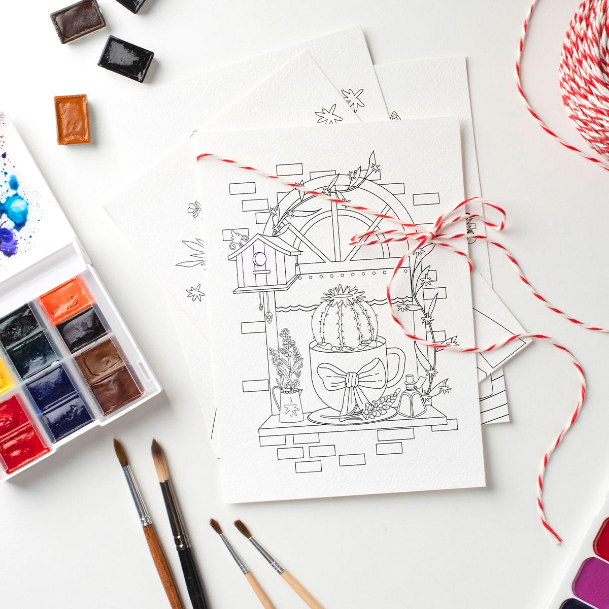 Coloring book page with a cactus design, watercolor paints, brushes, and string on a white surface.