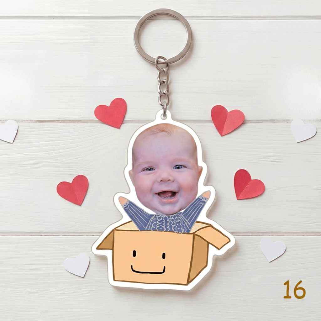Keychain with a smiling baby's face in a cardboard box surrounded by heart shapes on a wooden surface.
