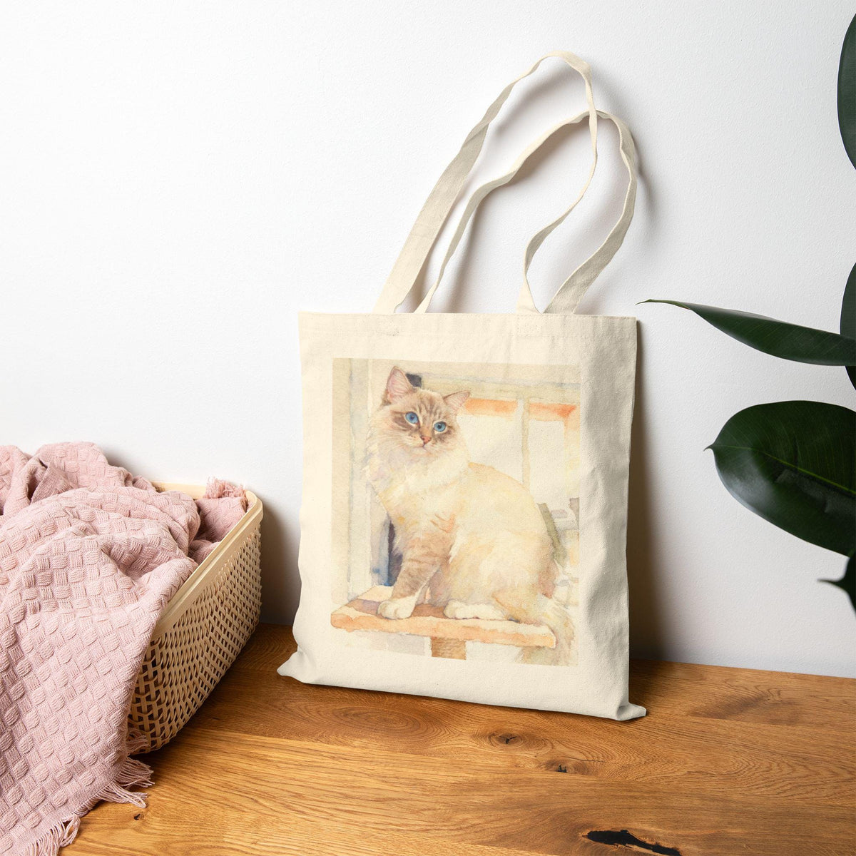 Tote bag with a cat design on a wooden surface next to a pink blanket and plant.