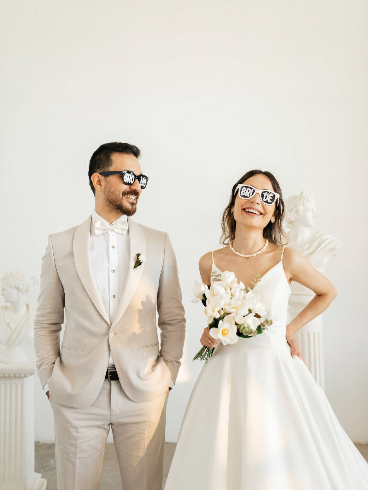 Man and woman in wedding attire with sunglasses on, standing against a white wall.